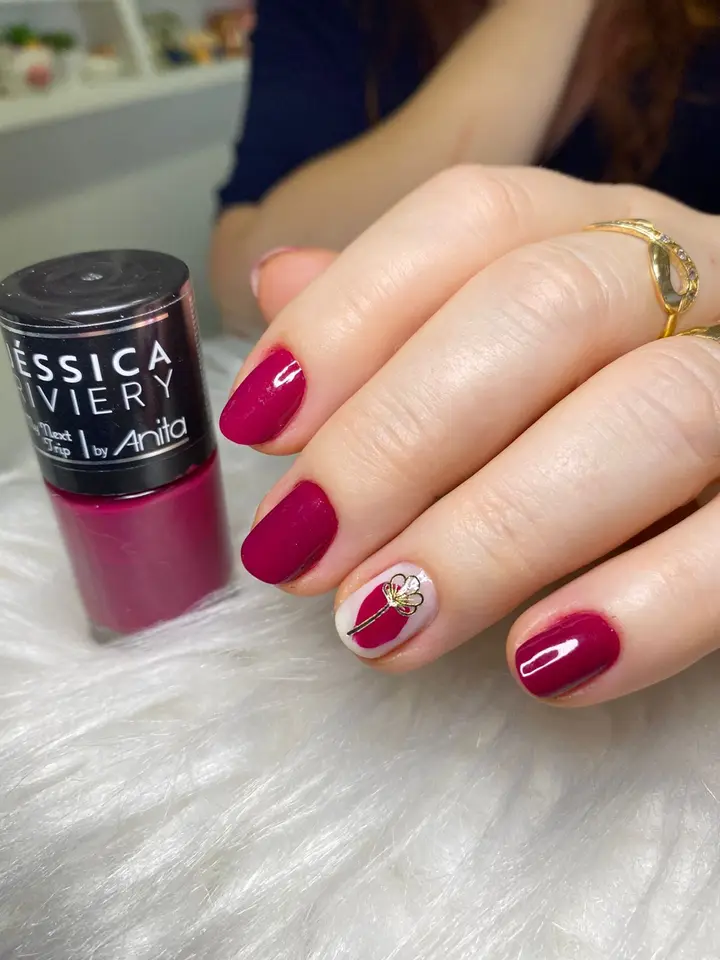 Unhas vinho com uma unha decorada com filete dourado, ao lado do vidro de esmalte